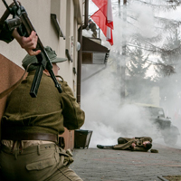 Inscenizacja historyczna: jeden żołnierz strzela z karabinu, drugi kuca pod budynkiem, trzeci leży na chodniku, w tle mgła i świerki.