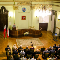 Wnętrze ozdobnej sali muzealnej, przy mównicy stoi mężczyzna, na sali siedzą na krzesłach ludzie, z sufitu zwisa żyrandol. 