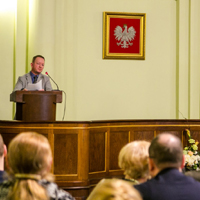 Publiczność zgromadzona w sali ratuszowej, stojący przed ludźmi mężczyzna mówiący do mikrofonu, na ścianie wisi oprawiony w ramkę wizerunek białego orła na czerwonym tle.