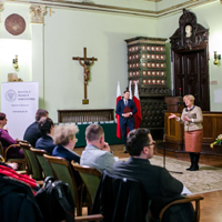 Zgromadzona na sali ratuszowej publiczność, mówiąca do mikrofonu kobieta, obok mężczyzna na tle biało-czerwonych flag, na ścianie wisi krzyż, w rogu sali piec kaflowy. 