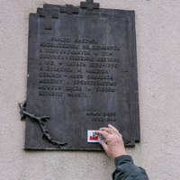 Mężczyzna naklejający naklejkę z logo Powstania Warszawskiego na wiszącą na ścianie budynku pamiątkową tablicę z napisami.