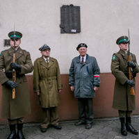 Czterech mężczyzn stojących pod ścianą budynku, na której wisi pamiątkowa tablica. Dwóch trzyma karabiny.