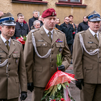 Trzech żołnierzy składa wiązankę kwiatów, z tyłu stojący pod ścianą ludzie.