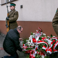 Mężczyzna w czarnej kurtce składa wiązankę biało-czerwonych kwiatów pod budynkiem, gdzie leżą już inne takie wiązanki, obok stoją żołnierze.