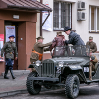 Dwóch mężczyzn trzyma innego mężczyznę wysiadającego z samochodu, na chodniku mężczyźni w mundurach, a nimi budynek z okratowanymi oknami. 