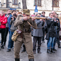 Mężczyzna w mundurze celuje z karabinu, w tle zgromadzona publiczność, niektórzy robią zdjęcia telefonami i aparatami, dalej budynki i drzewa. 