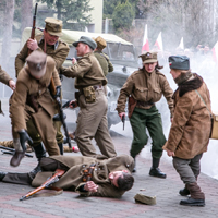 Inscenizacja historyczna: grupa osób w historycznych mundurach wojskowych w trakcie strzelaniny, w tle drzewa, biało-czerwone flagi i samochód.