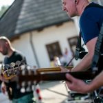 Trzech mężczyzn grających na gitarach, w tle budynki.