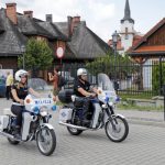 Kobieta i mężczyzna jadący ulicą na motocyklach , w tle widać drewniane zabudowania oraz biały budynek ratusza.