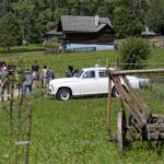 Biały zabytkowy samochód na trawniku, obok zgromadzeni ludzie, w tle drewniany budynek, z prawej strony drewniane ogrodzenie.