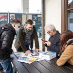 Trzech mężczyzn i jedna kobieta przy stoliku przed budynkiem, jeden z mężczyzn nachylony nad stołem, na którym rozłożone są materiały.