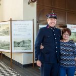 Mężczyzna w mundurze kolejowym stoi obok kobiety na terenie dworca kolejowego, z tyłu stelaże ze zdjęciami.