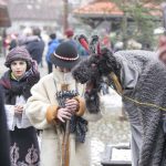 Postać w przebraniu turonia, obok dwóch chłopców w przebraniu pastuszków, w tle budynki oraz świerk.