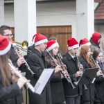 Dziewczynki i chłopcy grający na instrumentach muzycznych w orkiestrze, wszyscy mają na głowie czapki mikołajowe, w tle budynek.