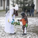 Dziewczynka przebrana za anioła podaje stojącemu naprzeciw chłopcu koszyk z zielonymi liśćmi. Z tyłu budynki i świerk.