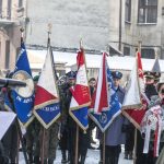 Mężczyźni w mundurach trzymający sztandary, z tyłu budynek kamienicy.