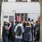 Grupa młodzieży zgromadzona na placu, wystające ponad ludzi drzewce sztandarów, na budynku wiszące tablice.