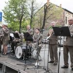 Zespół muzyczny w wojskowych mundurach stojący na scenie na ulicy, z tyłu drzewa i kamienica.