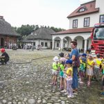 Grupa dzieci stojąca na brukowanym rynku miasteczka, przed nimi kobieta, obok czerwony wóz strażacki, w tle budynki.