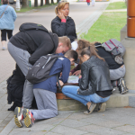 Grupka dzieci pochylona nad ławką na deptaku, obok nich kobieta, w tle aleja.