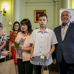 Mężczyzna w okularach podaje kobiecie papierową torbę, naprzeciwko niej kobieta trzymająca różę, obok inni ludzie.