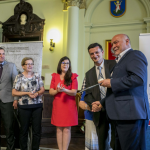 Uśmiechnięci mężczyźni i kobiety stojący na sali ratuszowej.