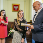 Starszy mężczyzna podaje dziewczynie dyplom, obok kobieta w sukience.