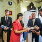 Kobieta podaje mężczyźnie dyplom, za nimi stoją ludzie.