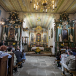 Wnętrze kościoła, po bokach siedzą ludzie w ławkach, naprzeciwko ozdobny ołtarz.