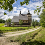 Drewniany kościół położony pośród zieleni, na tle nieba. Do kościoła biegnie polna droga.