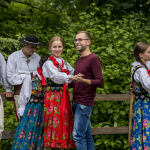 Kobieta w stroju góralki tańczy na scenie z mężczyzną w okularach, po bokach górale, z tyłu zieleń.