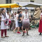 Kilka osób w strojach z epoki średniowiecza stoi na brukowanym dziedzińcu, w tle stoiska z rękodziełem.