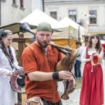 Mężczyzna w dawnym stroju grający na dudach, za nim kobieta grająca na tamburynie i kobieta grająca na bębenku, w tle namioty ze stoiskami z rękodziełem.