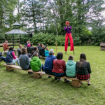 Clown na szczudłach chodzi po trawie, przed nim siedząca na ławkach publiczność. Z tyłu drzewa i altana.