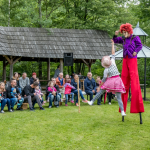 Clown bawiący się na trawie z małą dziewczynką, przed nimi siedząca na ławkach publiczność.