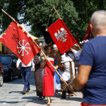 Grupka ludzi przebrana w stroje z epoki średniowiecza. Na przodzie kobieta niosąca czerwoną flagę.