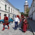 Mężczyzna w białej koszuli i niebieskich spodniach, po bokach dwóch mężczyzn w średniowiecznych strojach, z tyłu kamienice i kościół.