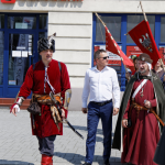 Mężczyzna w białej koszuli i niebieskich spodniach, po bokach dwóch mężczyzn w średniowiecznych strojach, z tyłu kamienica.