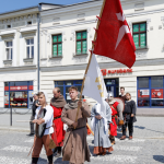 Grupka mężczyzn w średniowiecznych strojach, przechodzi przez ulicę, jeden z nich niesie czerwoną flagę, z tyłu kamienica.