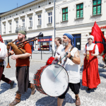 Grupa osób w średniowiecznych strojach przechodząca przez ulicę, niektórzy niosą instrumenty muzyczne, z tyłu kobieta w czerwonej sukni z tamburynem, niosąca czerwoną flagę. Z tyłu kamienice.