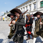 Kilku mężczyzn w dawnych strojach z epoki średniowiecza w kapeluszach na głowie przechodzą przez ulicę, z tyłu kamienice.