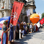 Grupa ludzi w dawnych strojach stojąca na środku rynku, duża czerwona flaga, po prawej stronie pomarańczowy balon.