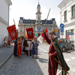 Grupa ludzi w dawnych strojach średniowiecznych, niektórzy trzymają czerwone flagi, z tyłu budynek ratusza, po bokach kamienice, na pierwszym planie mężczyzna w stroju rycerza.