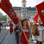 Dziewczyna z blond długimi włosami w średniowiecznym stroju, niesie czerwoną flagę, z tyłu inni ludzie, w tle budynek ratusza.