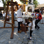 Dziewczynka wkładająca nóżkę do koszyka, obok mężczyzna trzymający koszyk na linach, obok drewniane umocnienia, z tyłu ludzie, w tle plenerowy ołtarz.