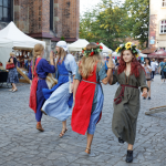 Dziewczęta z długimi włosami w średniowiecznych sukienkach tańczą na brukowanym dziedzińcu, obok widoczne mury kościoła oraz zgromadzeni ludzie.