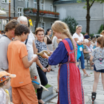 Kobieta w średniowiecznej sukni stoi obok grupy ludzi w strojach współczesnych, w tle ludzie i budynki.