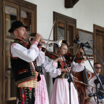 Kilkuosobowa kapela ludowa grająca przed drewnianym budynkiem, z tyłu inni ludzie.