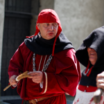 Mężczyzna w czerwonym płaszczu, łańcuchu na szyi nakryciu głowy, obok niego mężczyzna w stroju kata.