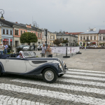 Zabytkowy, biało-granatowy samochód jadący ulicą, z tyłu kamienice, rynek i spacerujący po nim ludzie.